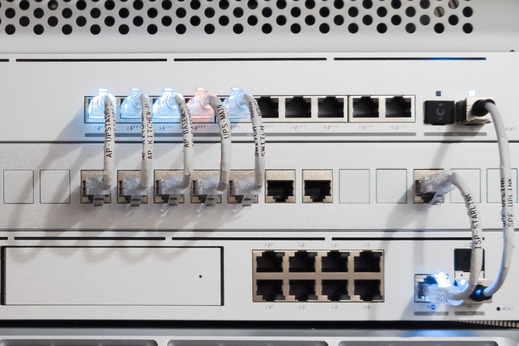 Close-up of short white patch cables between a switch and a patch panel, each with a printed label, among them AP-UPSTAIRS, AP-KITCHEN-01, AP-BASEMENT, UPS-MAIN, and ISP-STARLINK