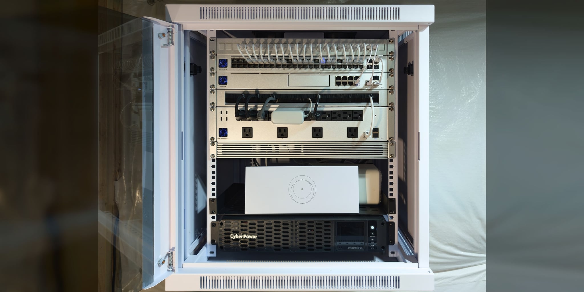 Commissioned wall-mounted UniFi rack behind a locking glass door — patch panel, PoE switching, UDM SE gateway, power distribution, and rack-mount UPS