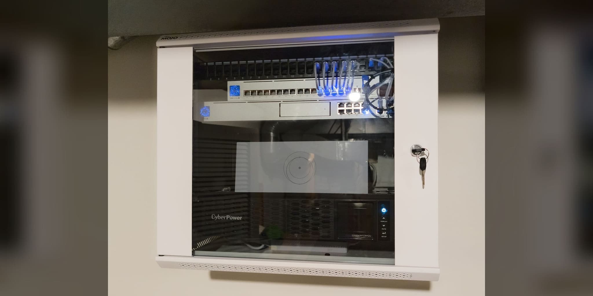 Wall-mounted network cabinet with patch panel, PoE switching, and a rack UPS behind a glass door (representative photo)