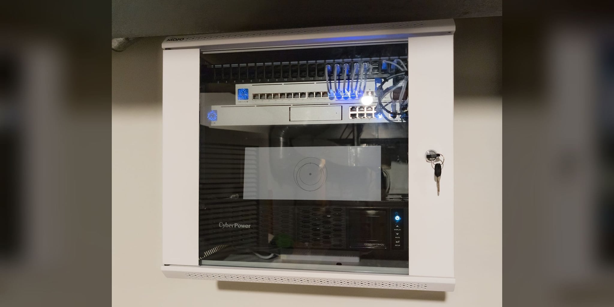 Wall-mounted network cabinet with patch panel, PoE switching, and a rack UPS behind a glass door (representative photo)