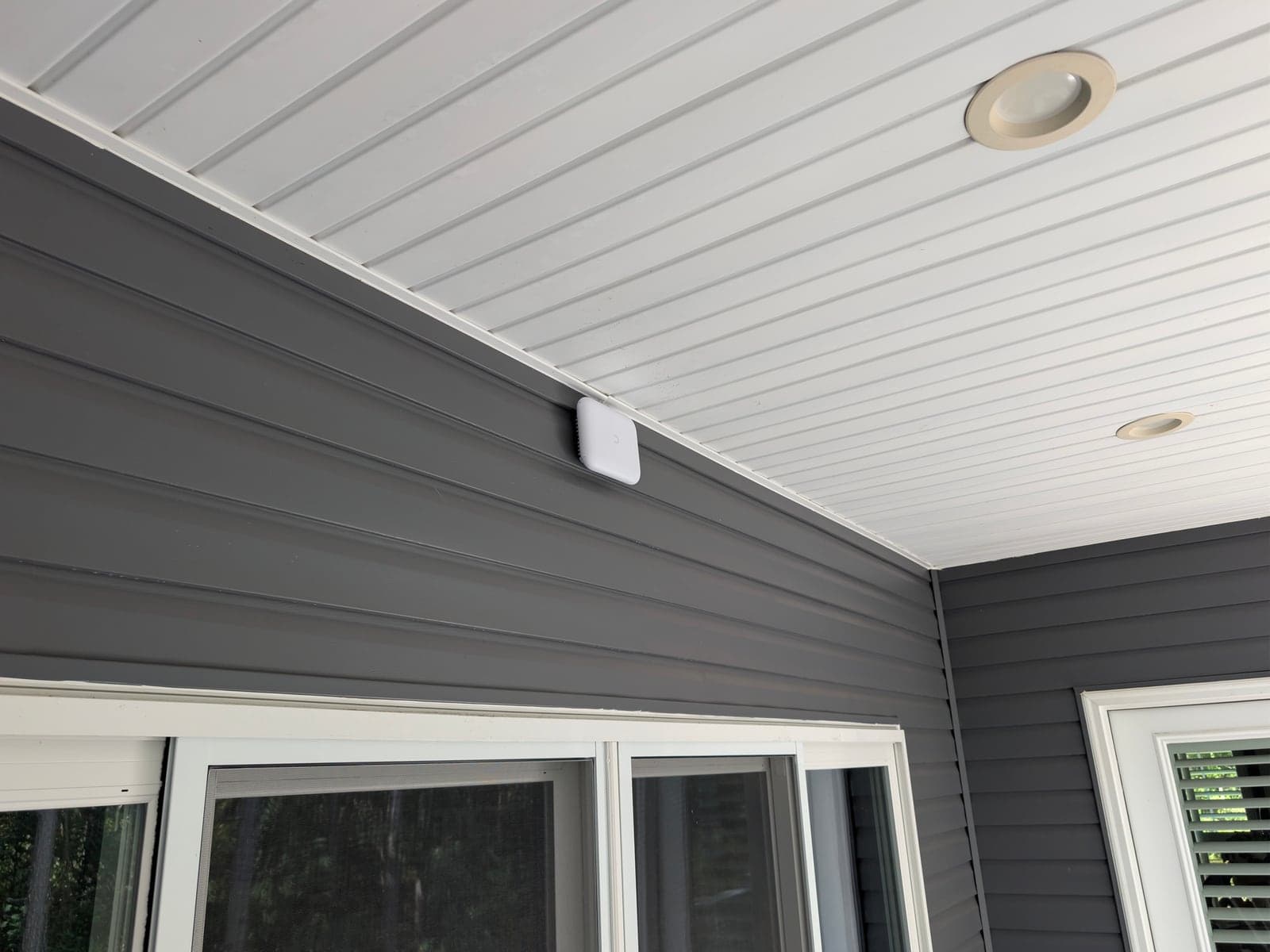 A white outdoor Wi-Fi access point mounted high on gray siding under a covered porch ceiling, above a row of windows