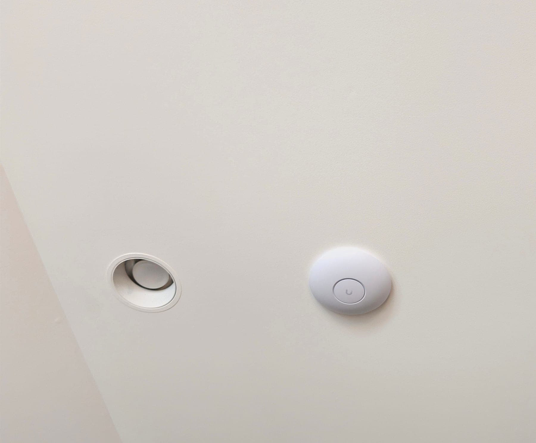 A round white ceiling access point set flush in a plain white ceiling, a short distance from a recessed light