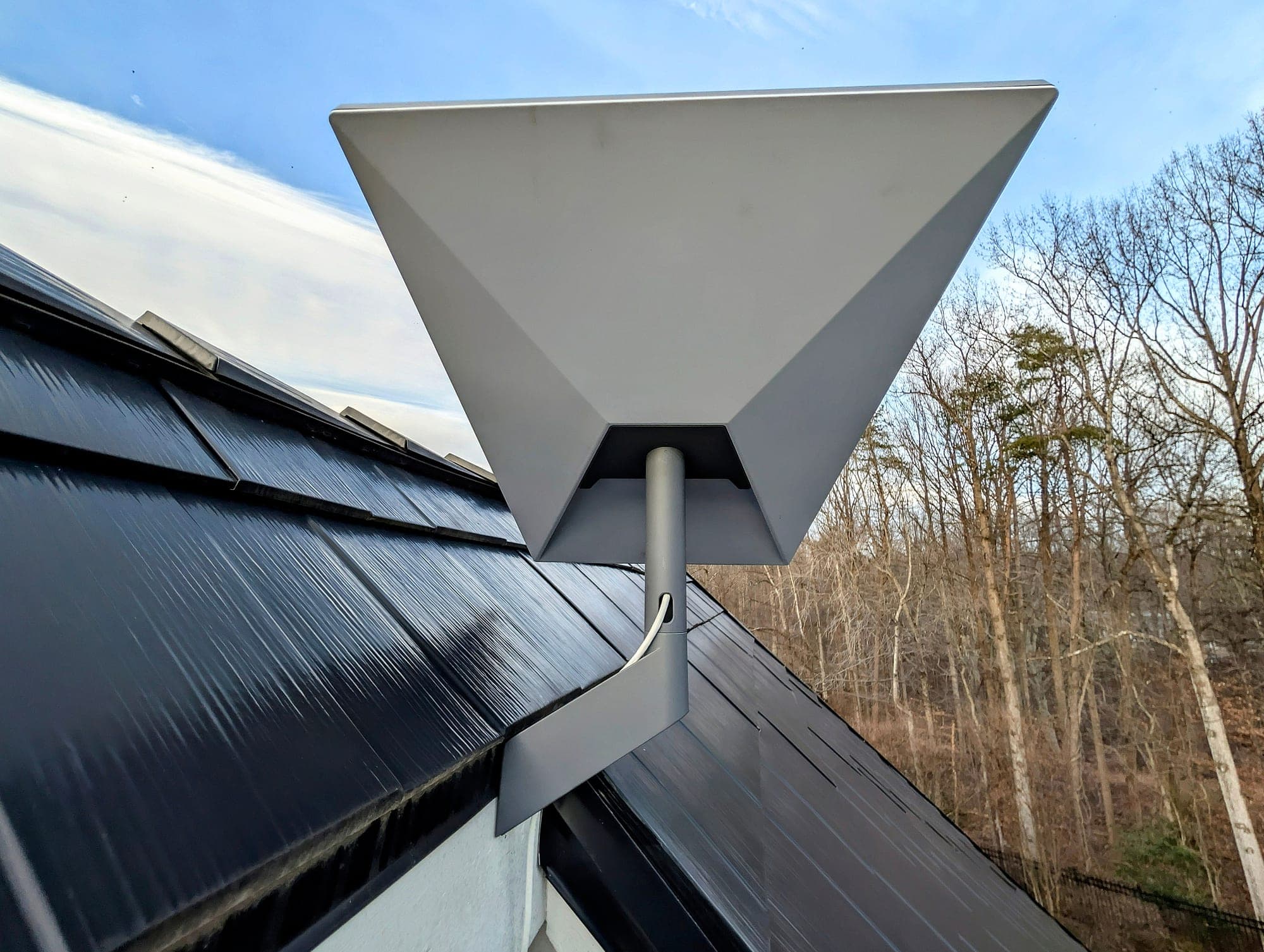 A Starlink dish on a gray bracket at the edge of a dark shingled roof, against a blue sky, with bare winter trees beyond